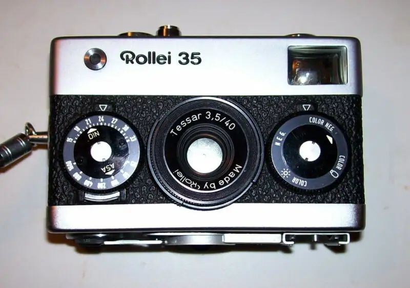 A vintage Rollei 35 film camera with a silver top, black textured front, and visible lens, dials, and viewfinder, displayed on a light surface.