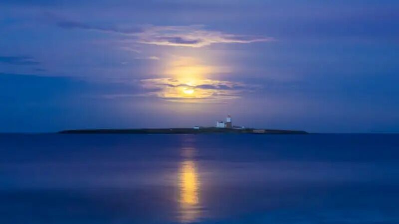 A small island with a lighthouse is silhouetted against a twilight sky. The moon glows above, casting a bright reflection on the calm, blue water surrounding the island.
