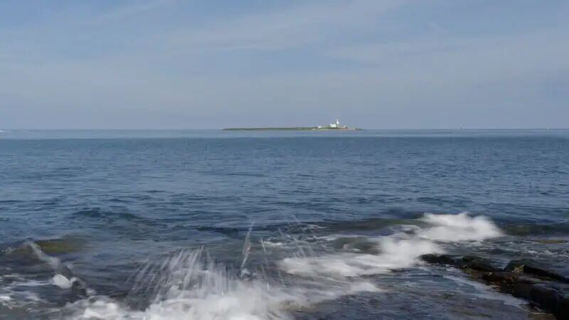 Waves splash against rocks in the foreground with a calm blue sea extending to the horizon; a small island with a lighthouse is visible in the distance under a clear sky.