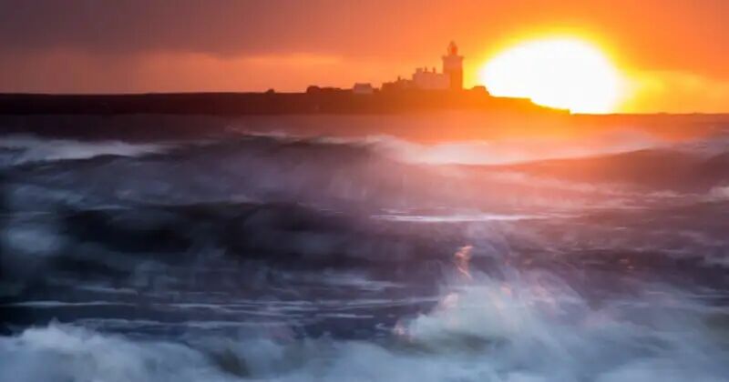 A dramatic sunset behind a distant lighthouse perched on a dark shoreline, with turbulent ocean waves in the foreground catching the light in streaks of blue, white, and orange.