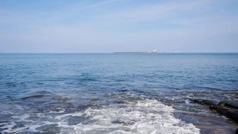 Waves gently crashing o<i></i>nto rocky shore in the foreground, with a small island and lighthouse visible in the distance under a blue sky.