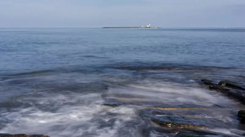 Waves gently wash over rocks in the foreground with calm blue sea stretching to the horizon. In the distance, a small island with a lighthouse is visible under a clear sky.